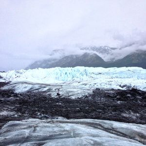 A serrated glacial wall, with dirty, gritty ice in the foreground and cloud covered mountains in the background. The photo almost appears black and white, aside from the teal blue of the seracs