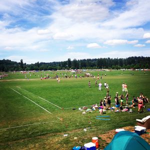 Green sports fields lined by trees under a cloudy blue sky. Tents surround the massive area with colourful athletes spotted throughout.